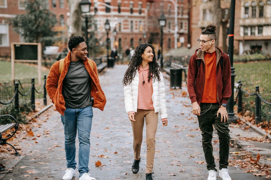 Positive young ethnic lady walking between multiethnic male friends with backpacks while spending free time together on autumn day in campus