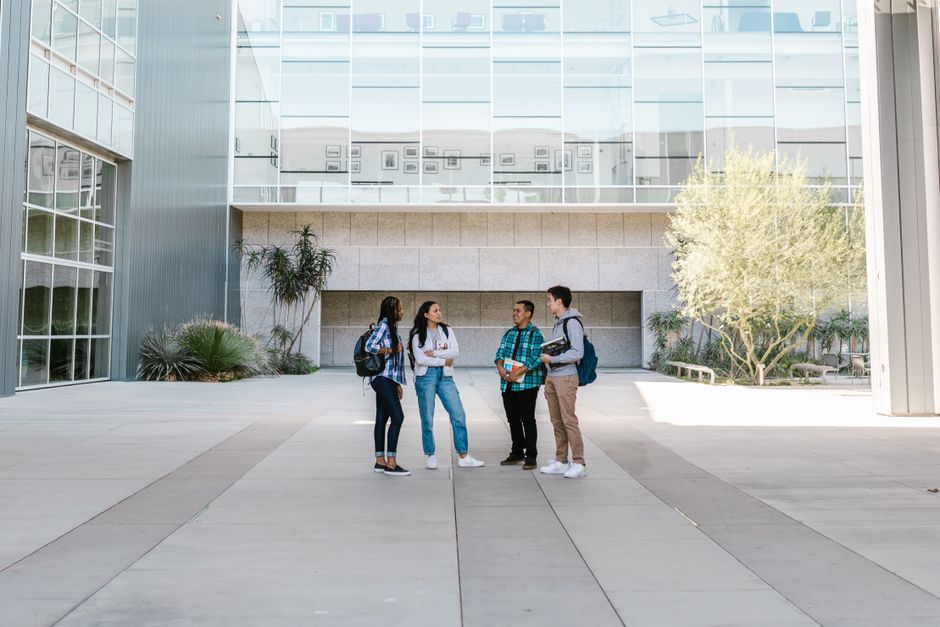 A group of diverse college students standing and talking in a sunny outdoor campus setting.