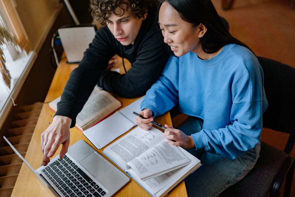 Two students collaborating on a study project using a laptop and textbooks on a wooden table.