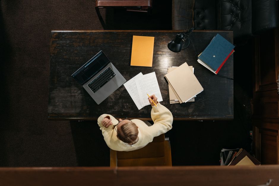 Person in yellow sweater working at a wooden desk with documents, folders, and a laptop.