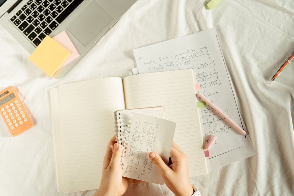 Hands holding a notebook with math equations, calculator, and laptop on a study desk.