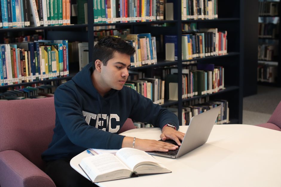 Student in library using laptop and textbooks for study and research.