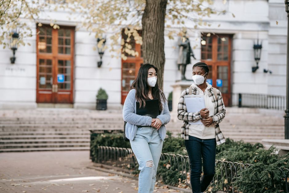 Focused young multiracial female students in casual clothes and protective masks chatting and carrying folders while strolling on campus sidewalk together