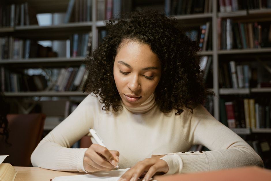 Young woman writing in a library, focusing on her studies with determination and concentration.