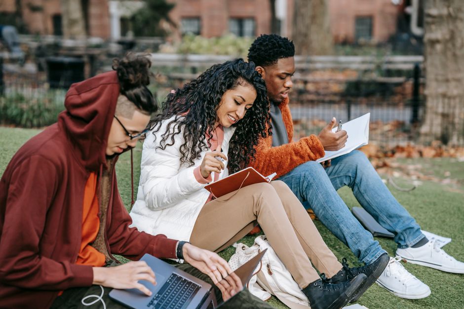 Group of focused multiracial classmates in casual outfits sitting on grass with paper and laptop while preparing for exams together