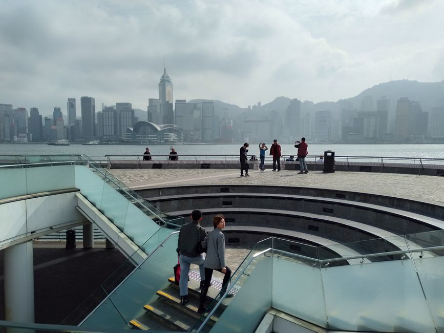 A foggy view of Hong Kong's Victoria Harbour from Kowloon, showcasing iconic skyline and people.