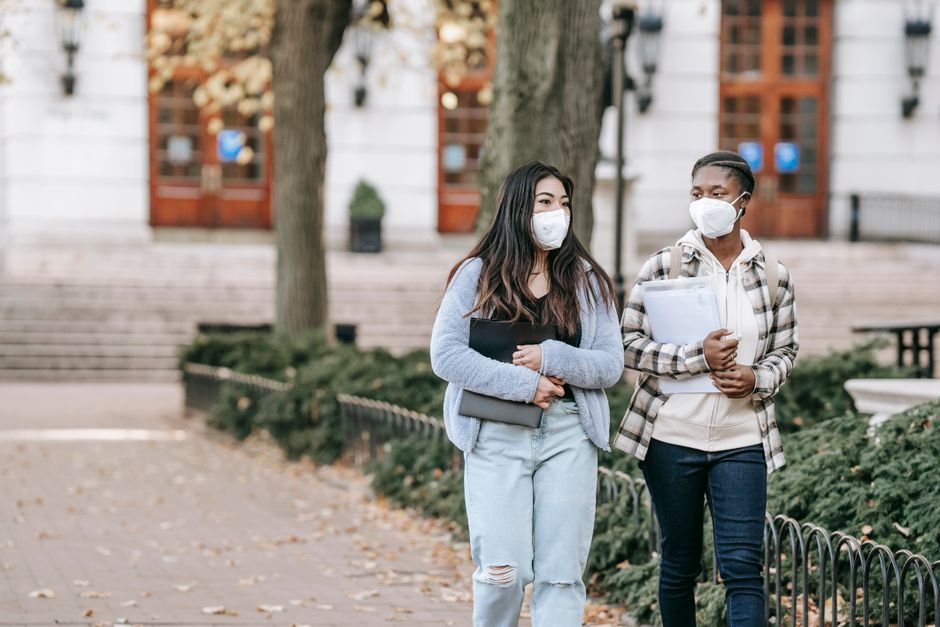 Calm young multiracial female students with folders wearing casual clothes and face masks strolling together on campus sidewalk