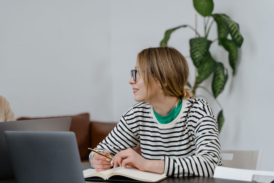 A woman in glasses sits at a desk, writing in a notebook with a laptop nearby, indoors.
